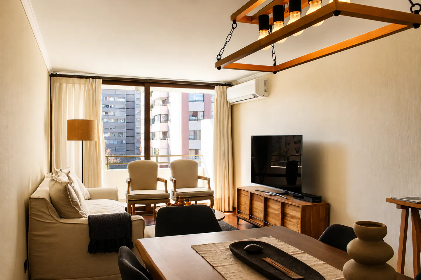 Living room with a sofa, chairs, TV, and dining table. A large window shows a city view. The room is lit by a wooden chandelier.