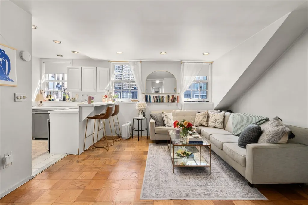 Bright open-concept living room with a beige sectional, glass coffee table, and kitchen island with two bar stools.