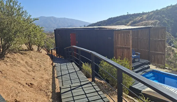 Exterior view of a modern black house with a red door, a wooden deck, and a blue pool on a hillside.