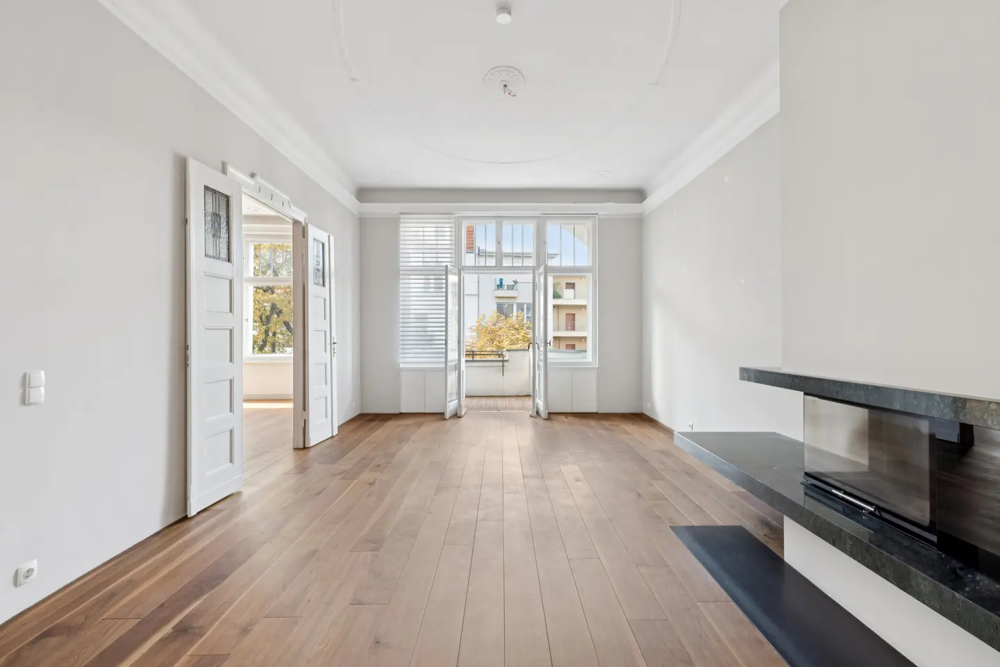 Bright, empty room with hardwood floors, white walls, and a black marble fireplace. Open doors lead to a balcony and another room.