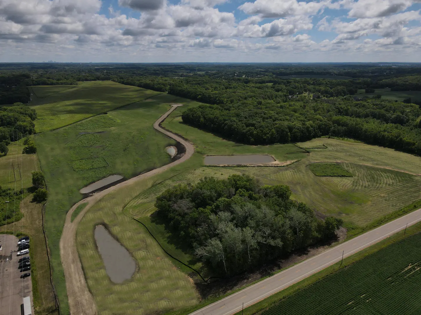Aerial view of a green field with ponds, trees, and a road under a cloudy sky.