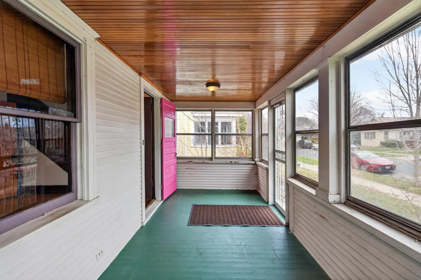 Enclosed porch with green floor, wood ceiling, and white walls. A pink door is open, and windows look out to a street.