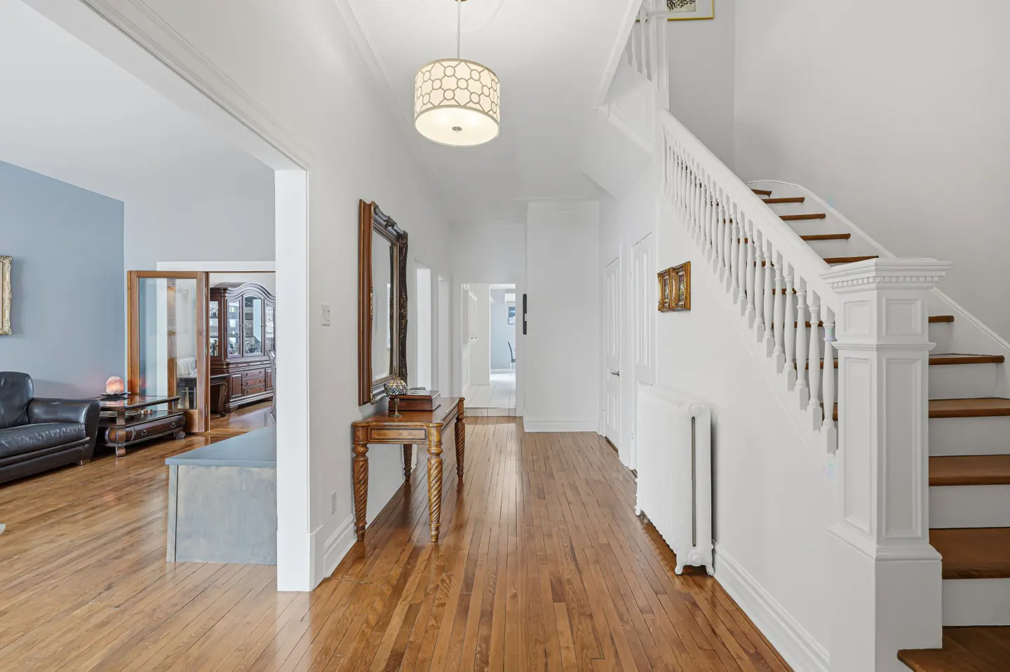 Bright hallway with hardwood floors, white walls, and a staircase. A wooden table with a mirror stands to the left. A living room is visible through an archway.