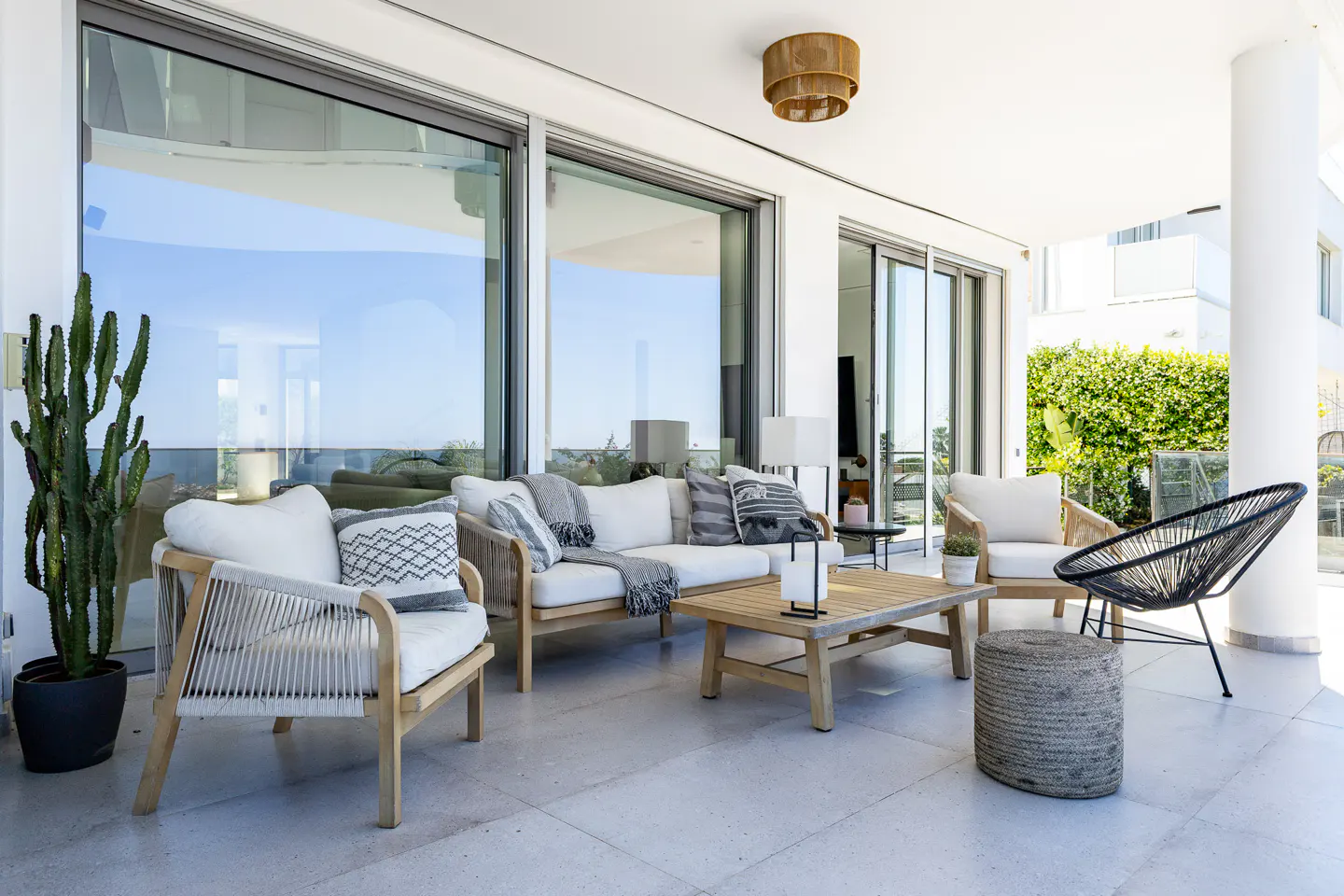 Outdoor patio with white sofa, wooden chairs, and coffee table. Large windows reflect blue sky. Cactus in pot on the left.
