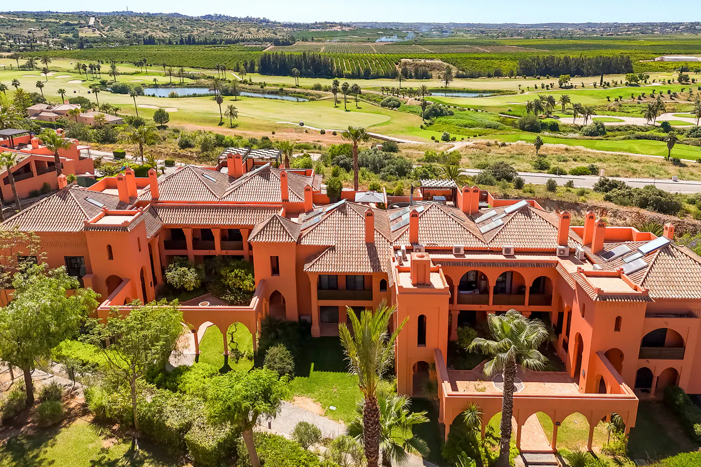 Aerial view of a terracotta-colored resort with a tile roof, surrounded by green lawns, palm trees, and a golf course in the background.