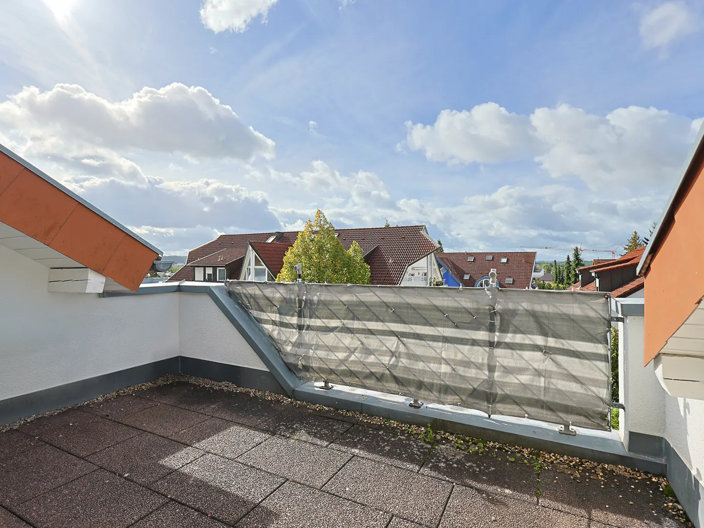 Rooftop terrace with brown tiles, white walls, and a gray privacy screen. Houses with red roofs are visible in the background under a cloudy sky.