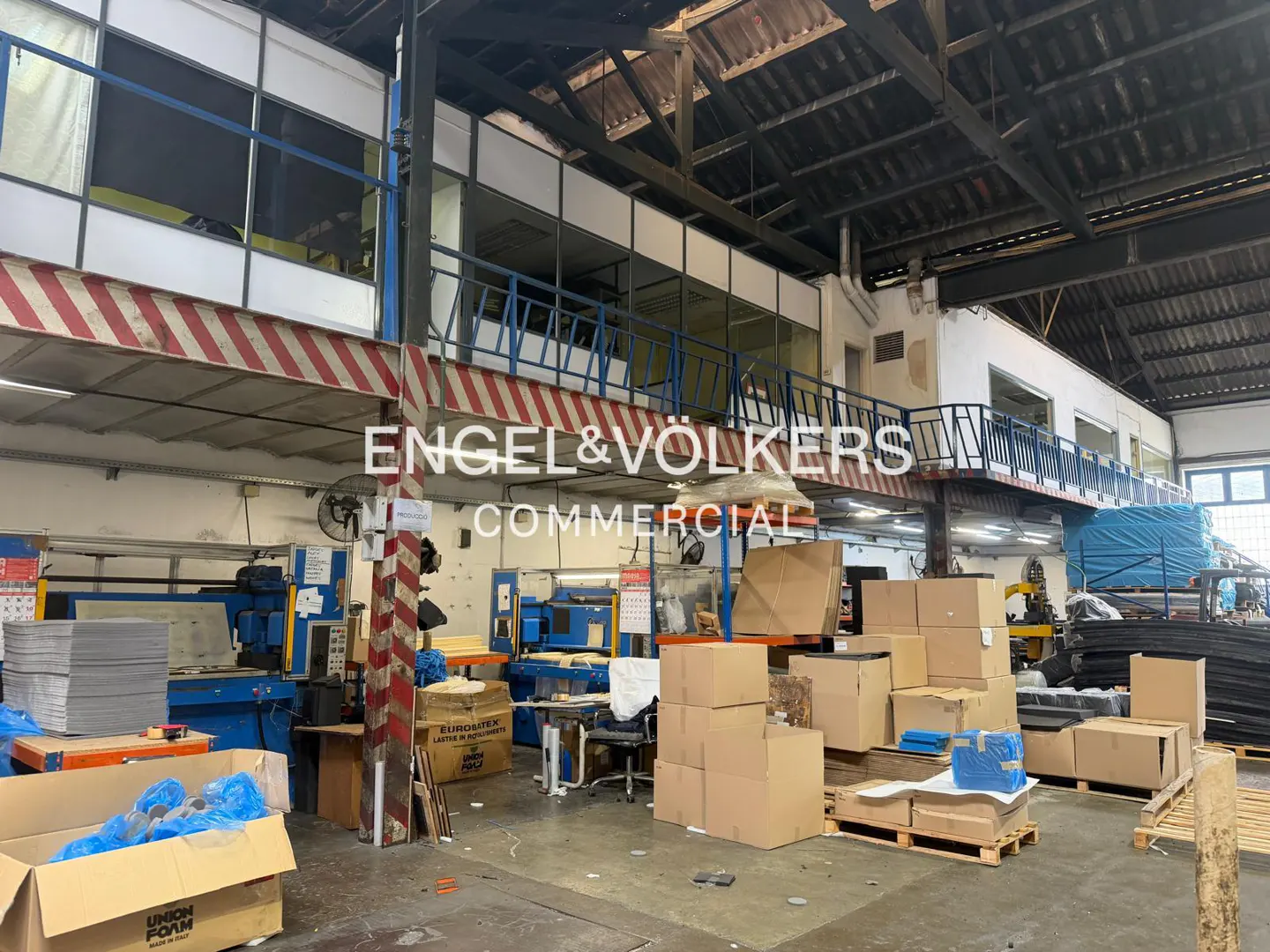 Interior view of a commercial warehouse with machinery, boxes, and a mezzanine level. The space is lit with natural light.