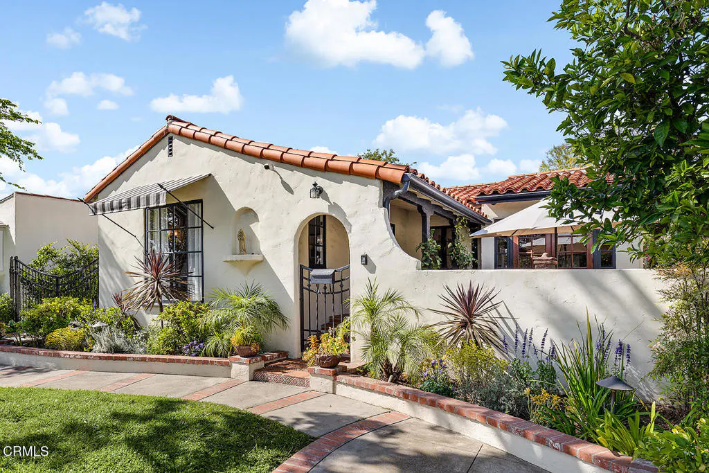 Exterior view of a cream-colored house with a red tile roof, black framed windows, and a black metal gate. The house is surrounded by lush greenery and a blue sky with white clouds.
