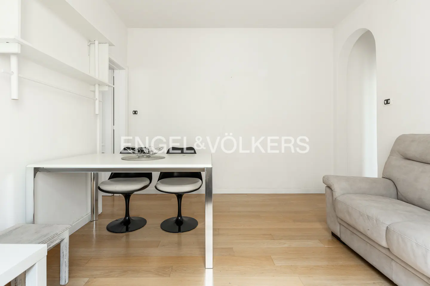 Bright, minimalist apartment interior with white walls, light wood floors, a white table with two chairs, and a gray sofa.
