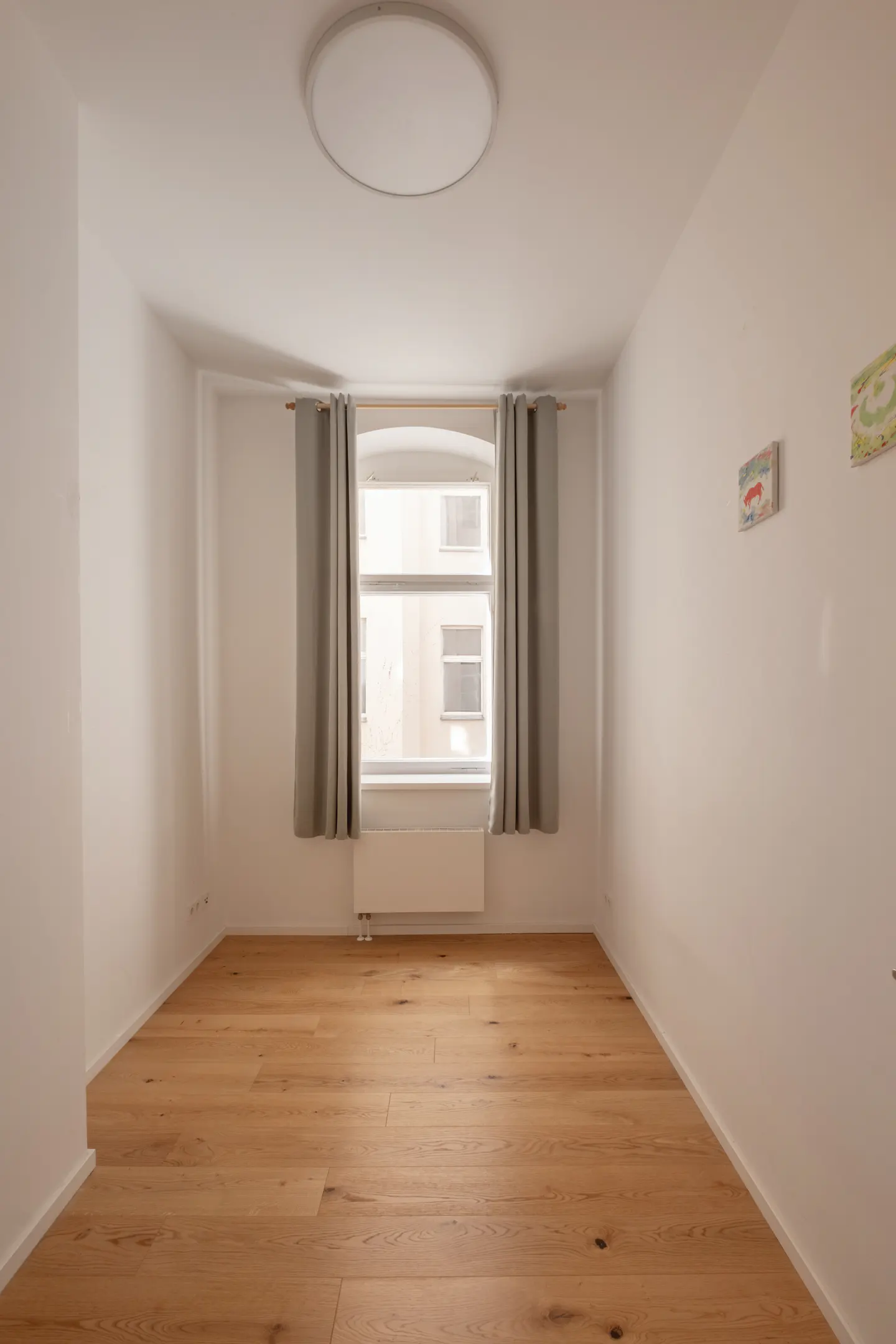 Empty room with white walls, light wood flooring, a central window with grey curtains, and a round ceiling light.