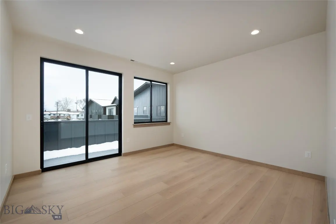 An empty room with light wood floors, white walls, and black-framed windows. Outside, snow covers the ground and nearby buildings.