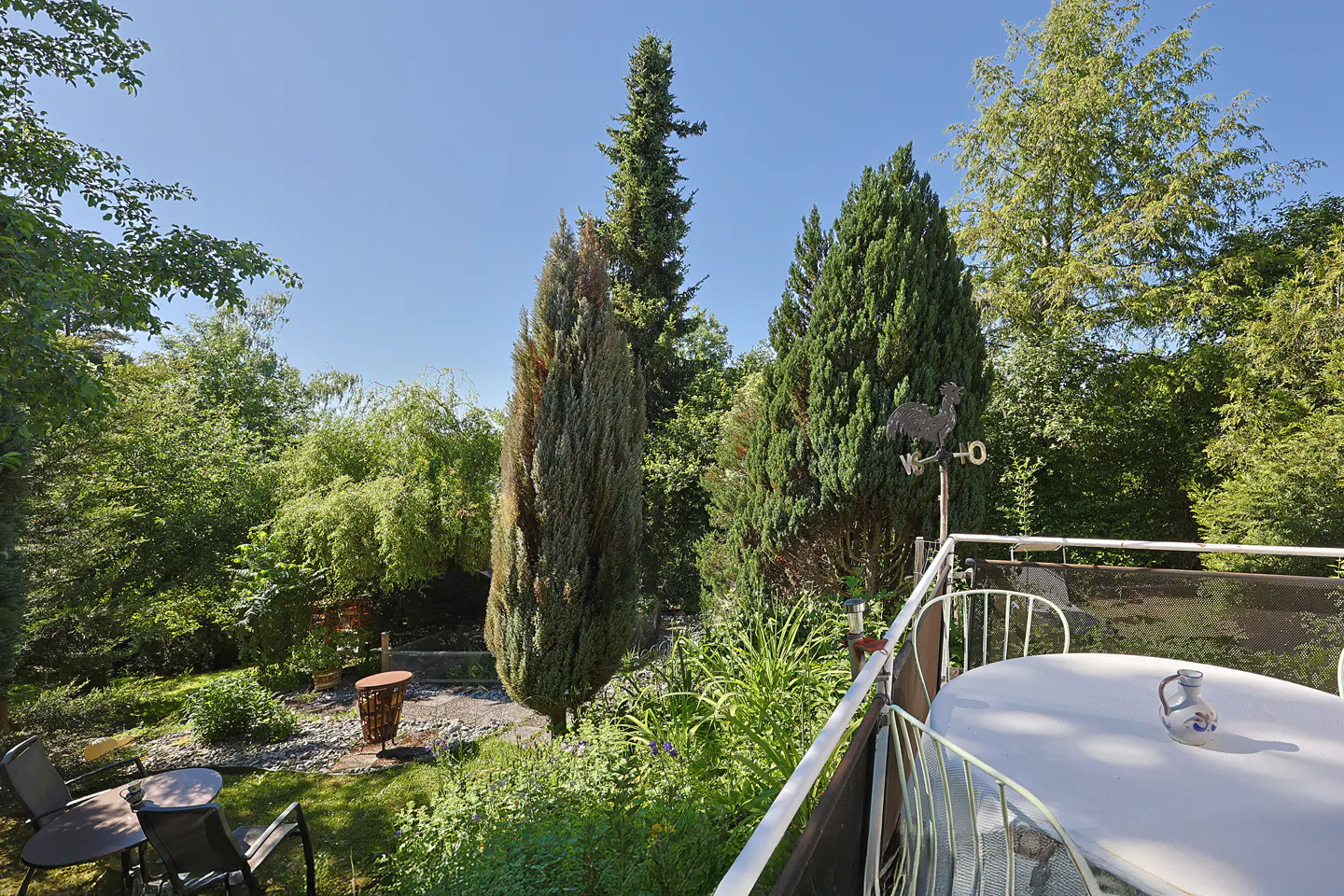 A sunny balcony with a white table overlooks a lush green garden with tall evergreen trees and a rooster weather vane.