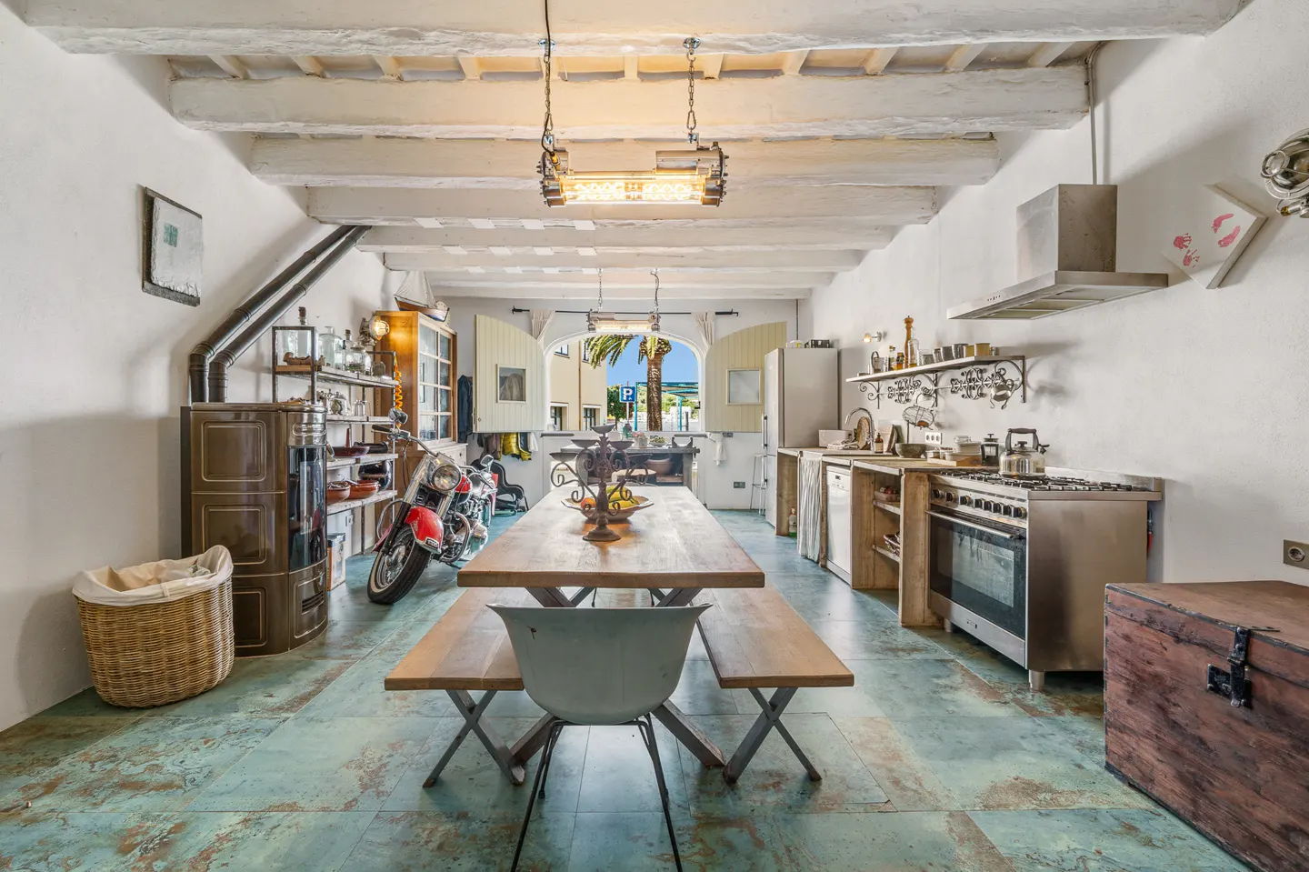 A bright, eclectic kitchen with a long wooden table, benches, and a motorcycle parked inside. White walls and ceiling beams add to the spacious feel.