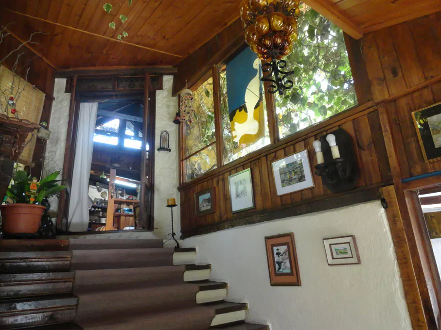 Interior view of a home's entryway with carpeted stairs, wood paneling, and framed art on white walls. A stained glass window and amber chandelier add character.