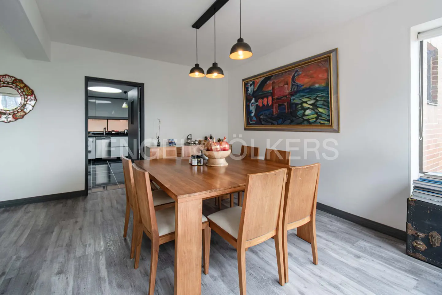 A dining room with a wooden table and chairs, a painting on the wall, and a view into the kitchen.