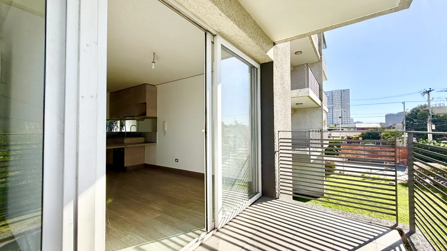 Open sliding glass doors lead from a balcony with a metal railing into a modern apartment with wood floors and a kitchen.