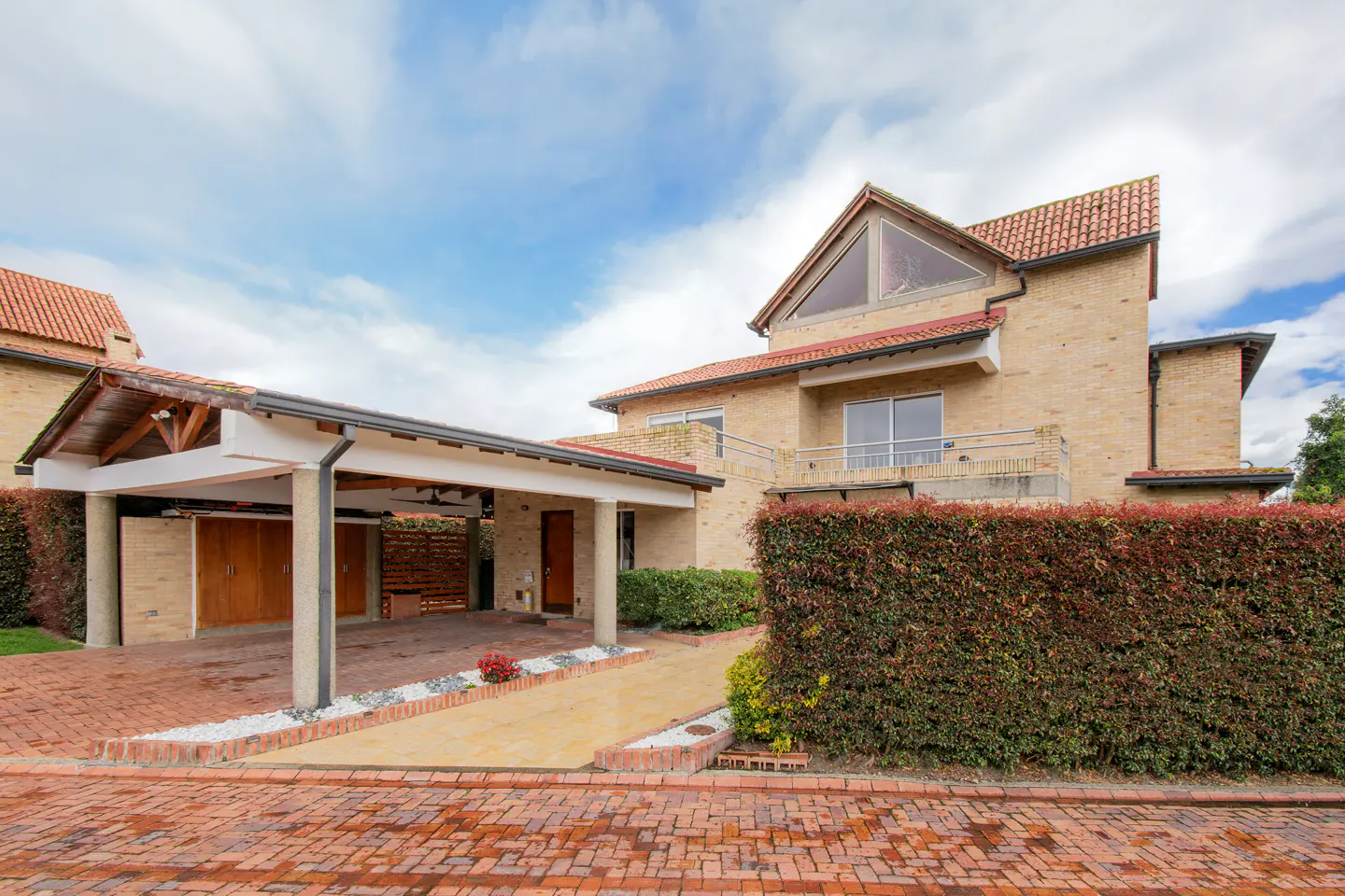 Two-story tan brick house with a red tile roof, a large carport, and a tall privacy hedge under a blue cloudy sky.