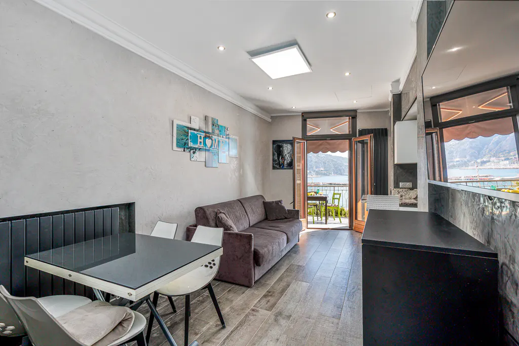Living room with a brown sofa, a black table with white chairs, and a balcony overlooking a lake and mountains.