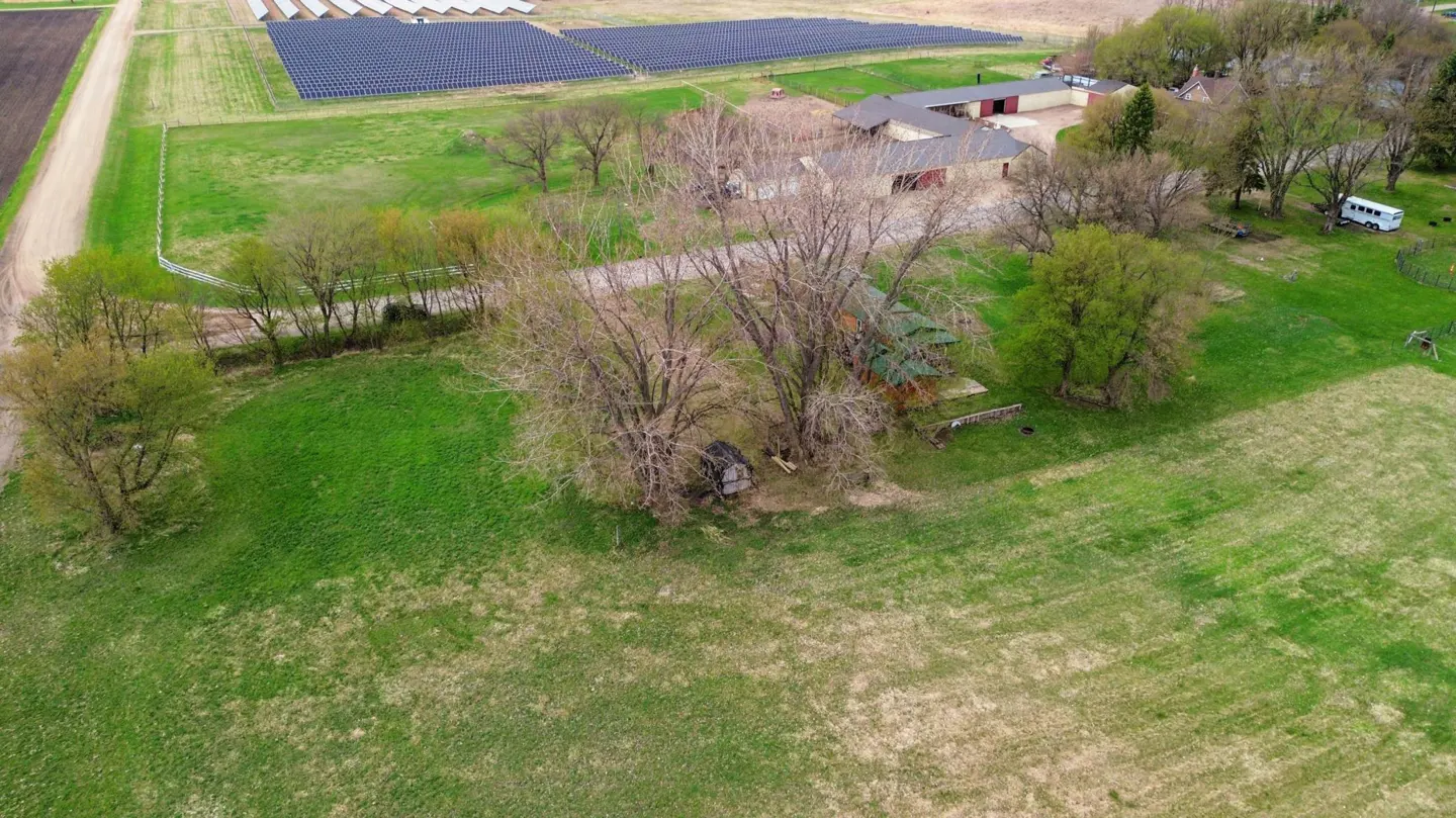 Aerial view of a rural property with green fields, trees, buildings, and a solar panel array in the background.