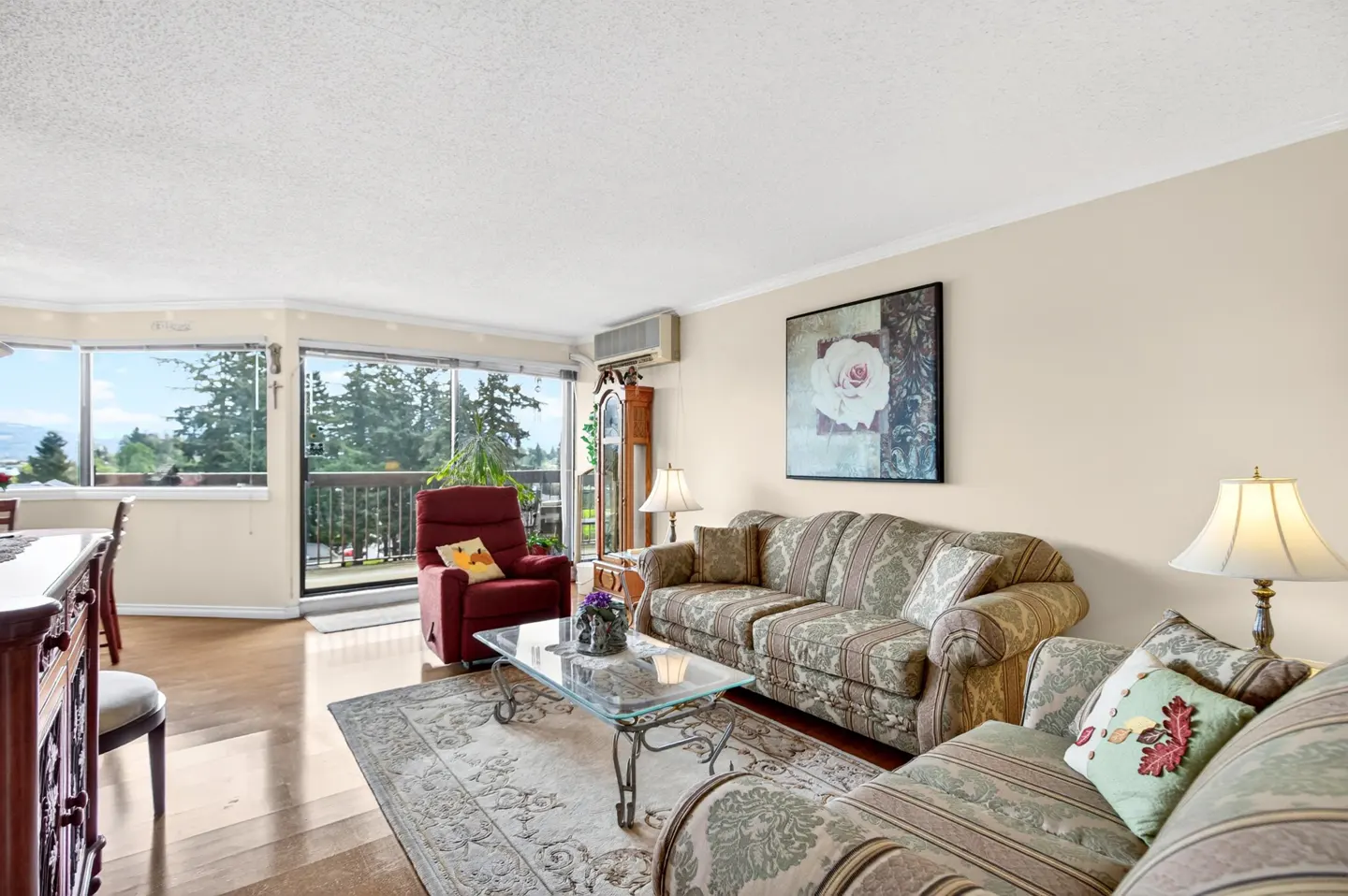 Living room with hardwood floors, patterned sofas, glass coffee table, and a red recliner near a balcony with a view.