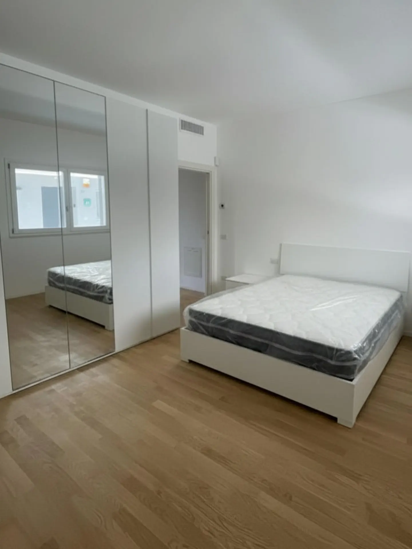 Bright bedroom with wood floors, white walls, and a bed with a mattress still in plastic. Mirrored closet doors reflect the room.