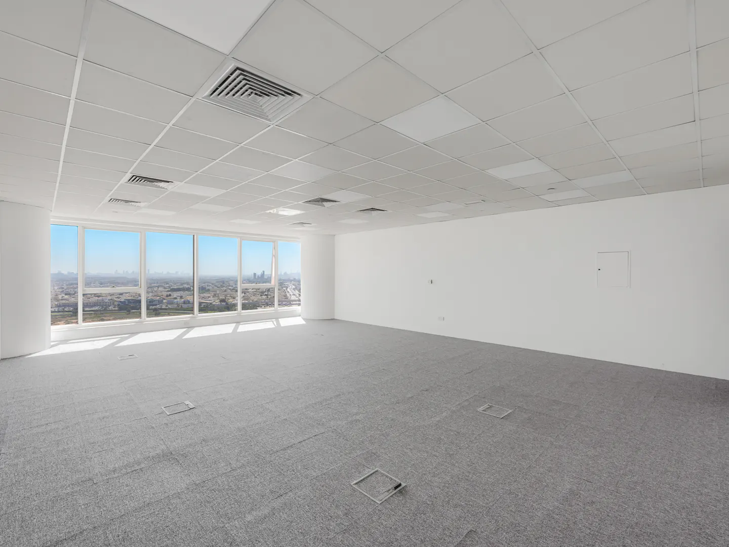 Bright, empty office space with gray carpet, white walls, and large windows overlooking a city skyline.