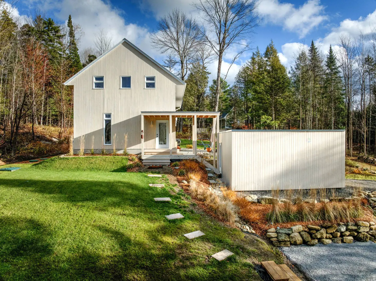 Modern light-colored house with a porch and a detached garage, surrounded by green lawn and trees under a blue sky.