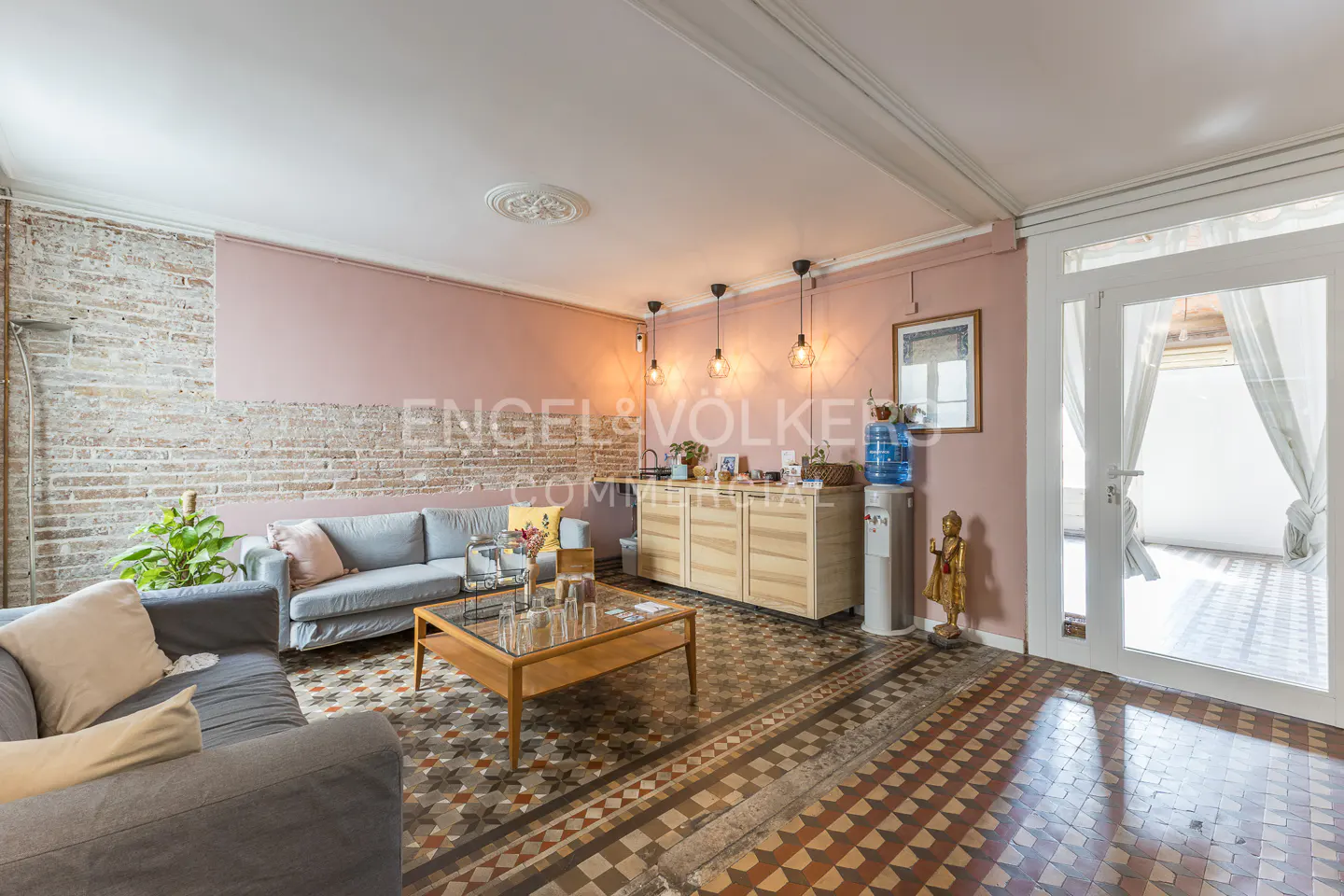 Living room with gray sofas, wood table, and patterned tile floor. Exposed brick wall and pink accent wall. White double doors lead to a bright space.