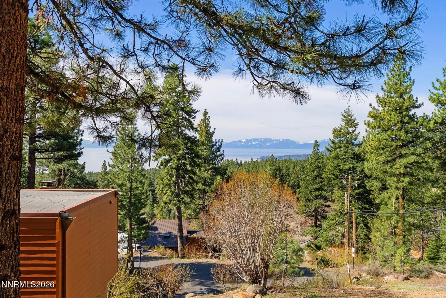 View of Lake Tahoe and mountains from a wooded property. Pine trees frame the scene. A building is visible on the left.