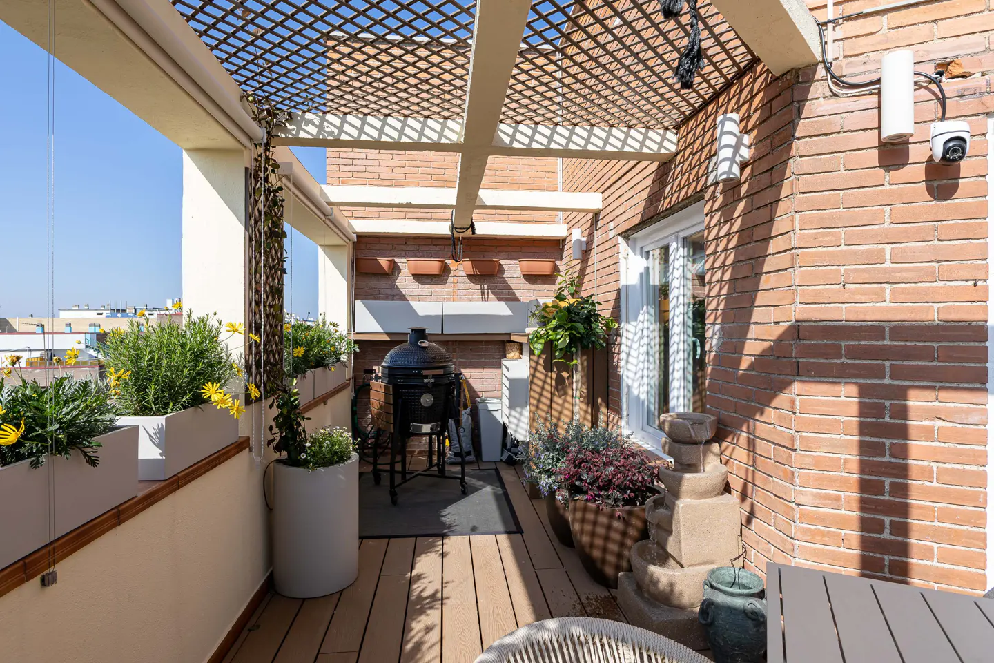 A rooftop patio with a grill, plants in pots, and a lattice pergola. The patio has a brick wall and a wooden floor.