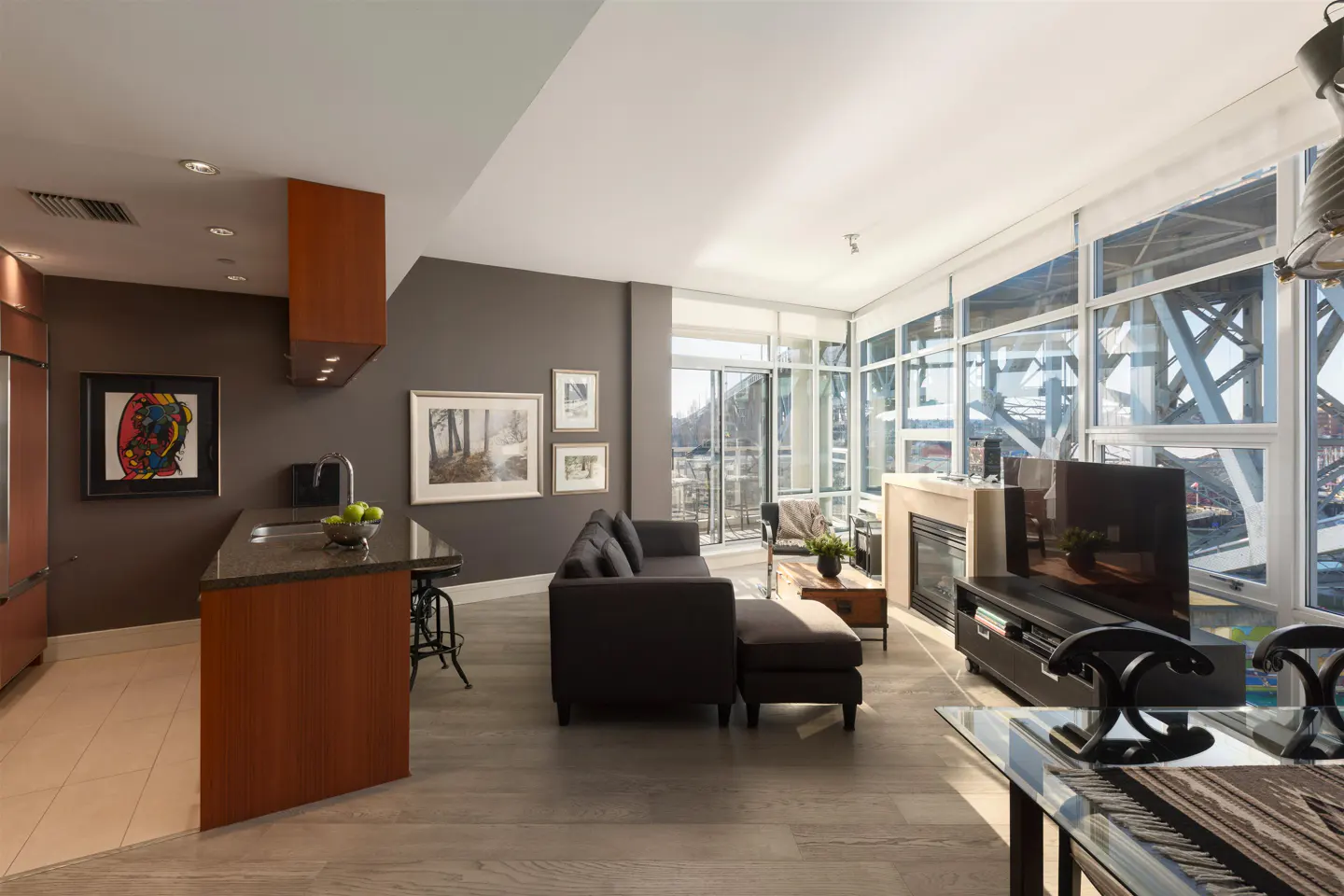 Bright condo interior with a gray sofa, wood floors, and a kitchen island. Large windows offer a view of a bridge.