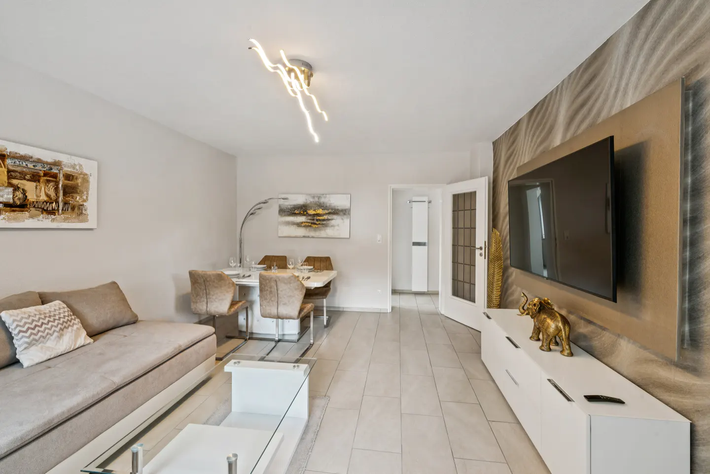 A modern living and dining area with a beige sofa, white table, and a TV on a patterned wall.