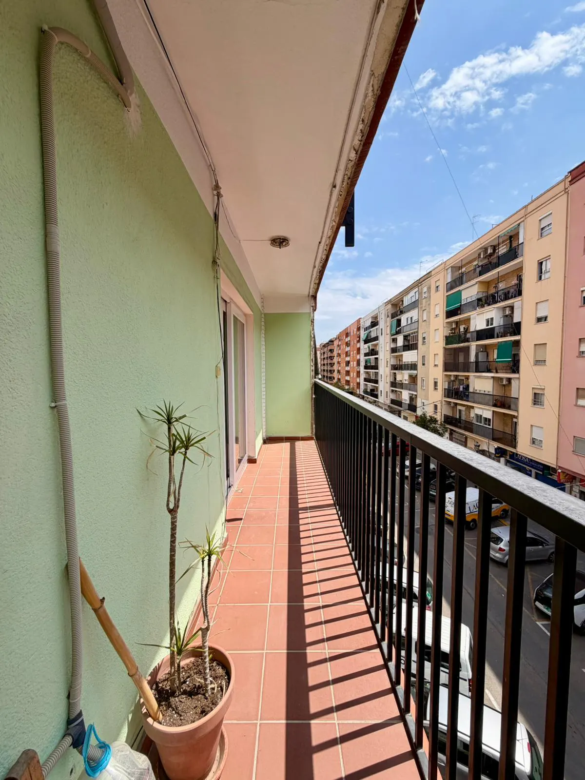 A balcony with a potted plant, red tile floor, and black railing overlooks a city street with colorful buildings.
