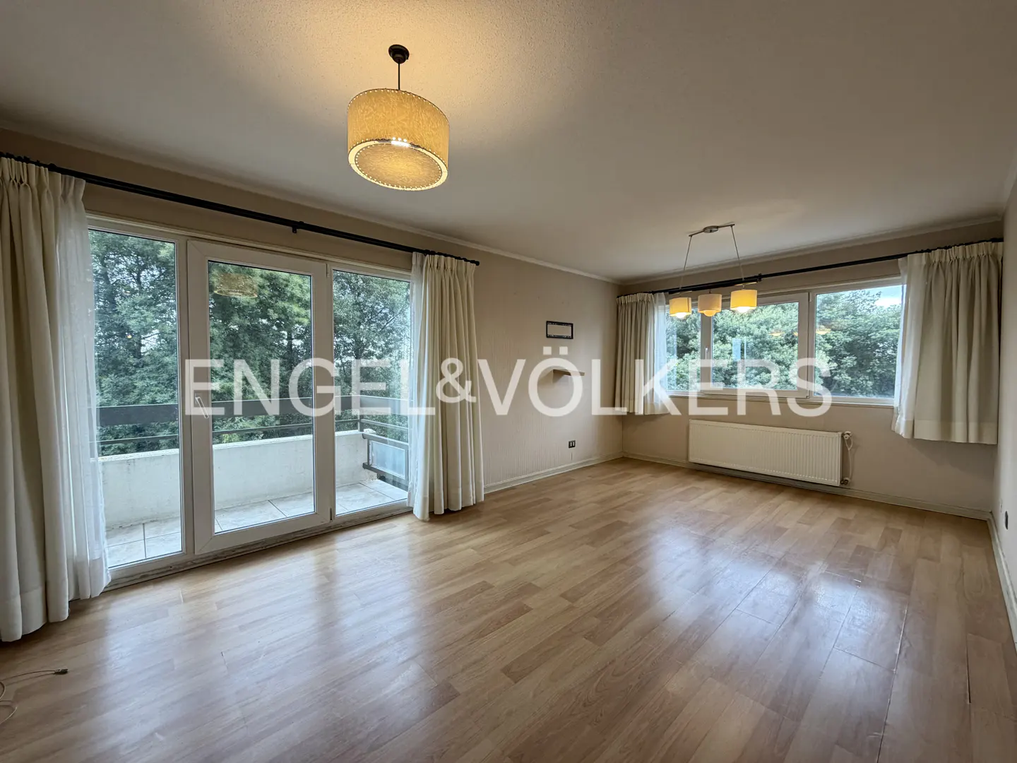 Bright, empty living room with hardwood floors, beige walls, and white curtains. Balcony access and large windows offer a view of trees. Engel & Völkers logo visible.