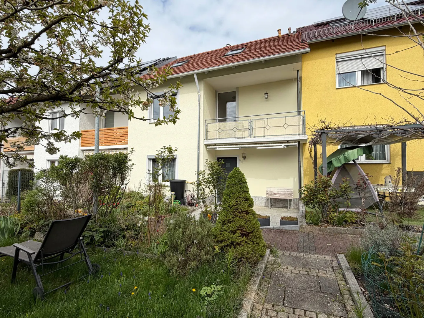 Exterior view of a two-story house with a balcony and a garden with a chair and plants.