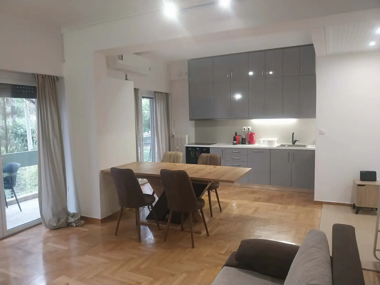 A bright, open-concept apartment with a dining table, grey cabinets, and a balcony with a view of trees.