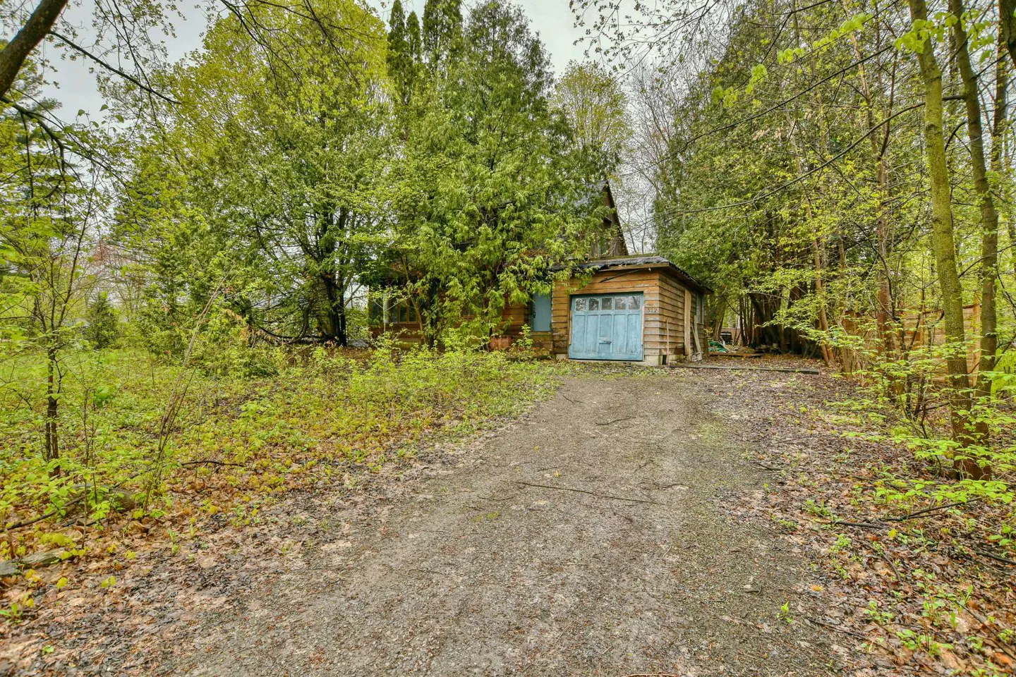Exterior view of a small, weathered house with a blue garage door, surrounded by lush green trees and a gravel driveway.