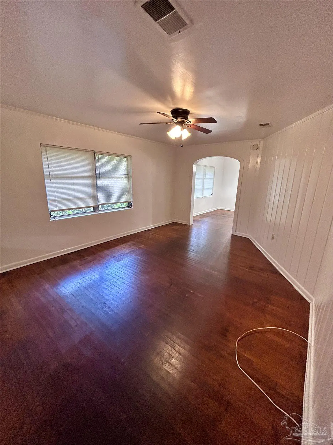 Empty living room with dark hardwood floors, white walls, a ceiling fan, and an arched doorway to another room.
