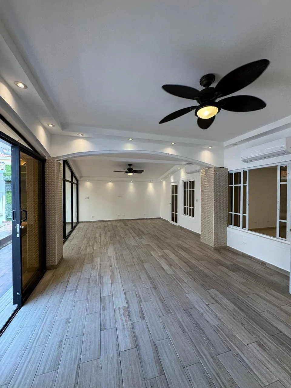 Bright, empty room with gray wood-look tile, white walls, and black ceiling fans. Sliding glass doors lead to the outside.