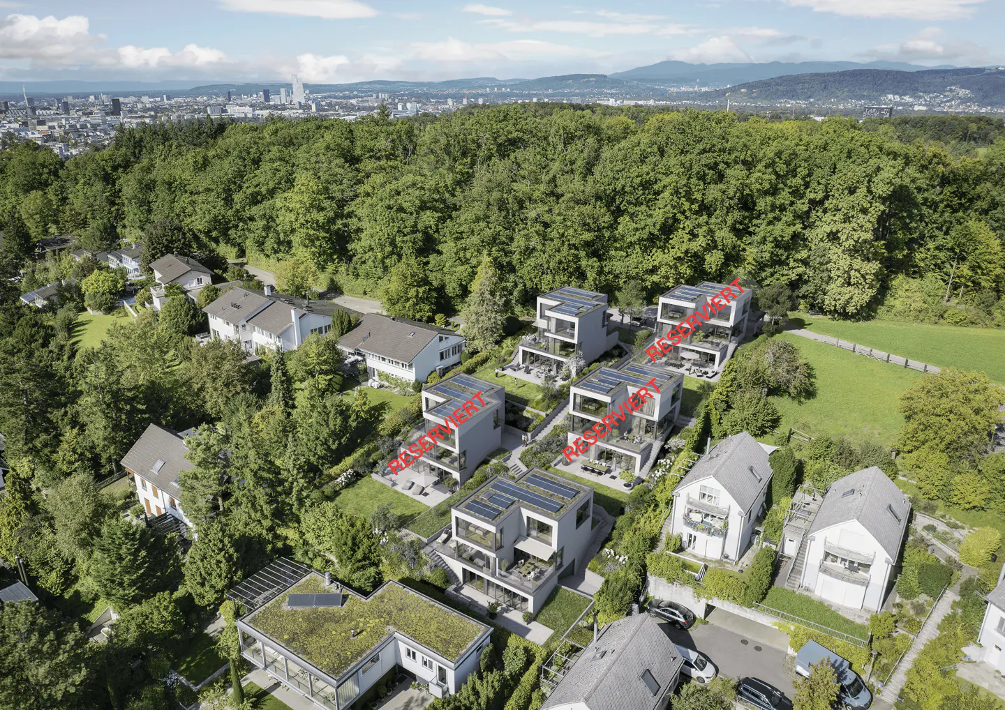 Aerial view of modern, white, multi-story homes with solar panels, nestled in a lush, green, wooded area with a city skyline in the distance.