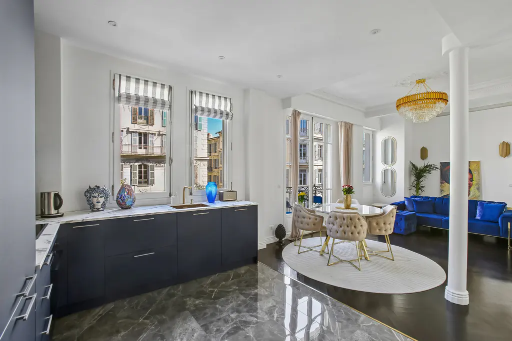 Bright, open-concept apartment with a kitchen, dining area, and living room. Blue cabinets, marble floors, and a blue velvet sofa are visible.