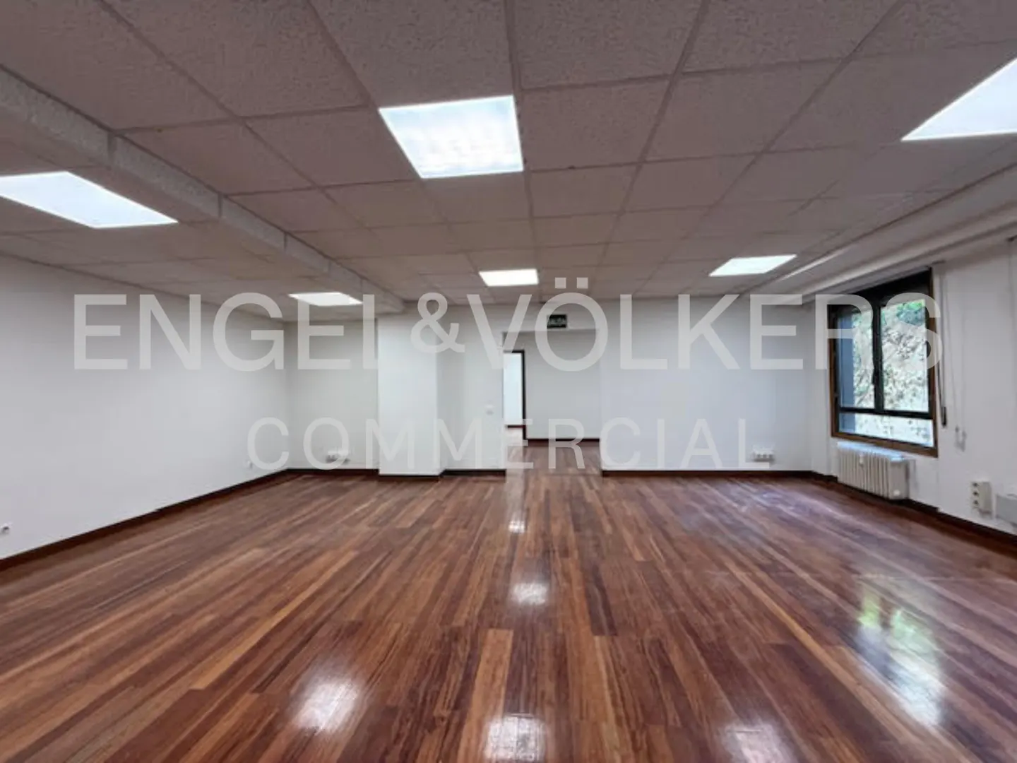 Empty commercial space with wood floors, white walls, and a window. Ceiling lights illuminate the room.