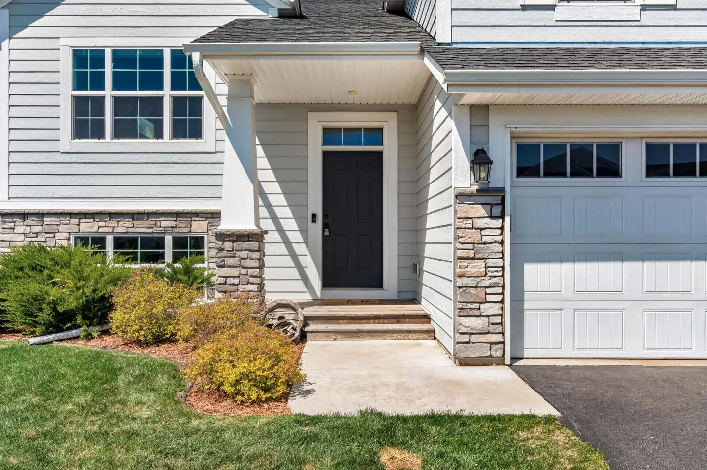 Exterior of a two-story house with light gray siding, stone accents, and a black front door. A white garage door is visible on the right.