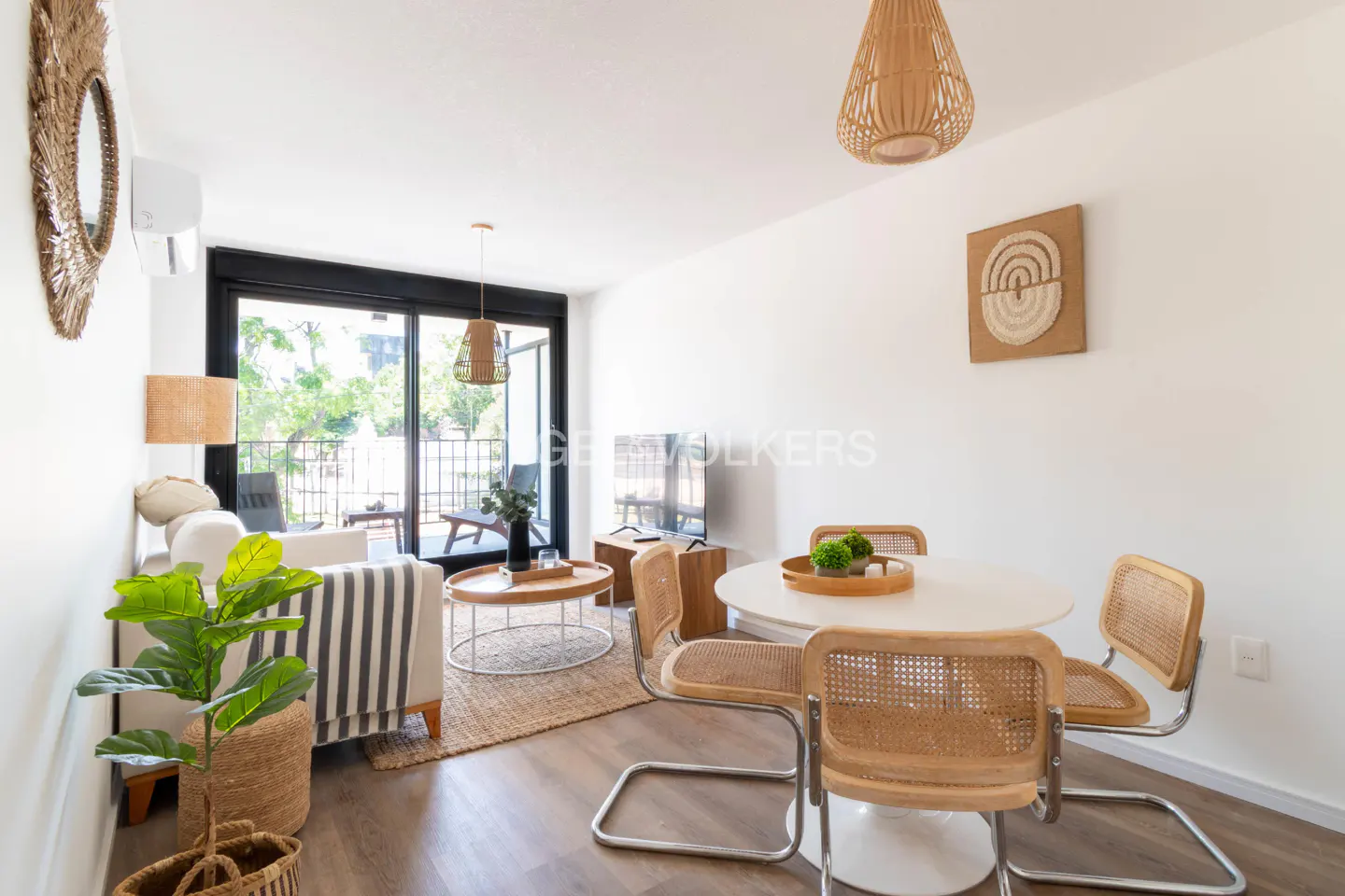 Bright living room with white walls, wood floors, and a round white table with four wicker chairs. A striped couch and a plant are visible.