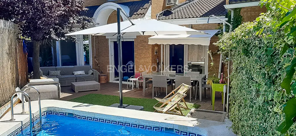 Outdoor patio with a blue pool, gray sofa, table, chairs, and a large white umbrella. Green plants surround the area.