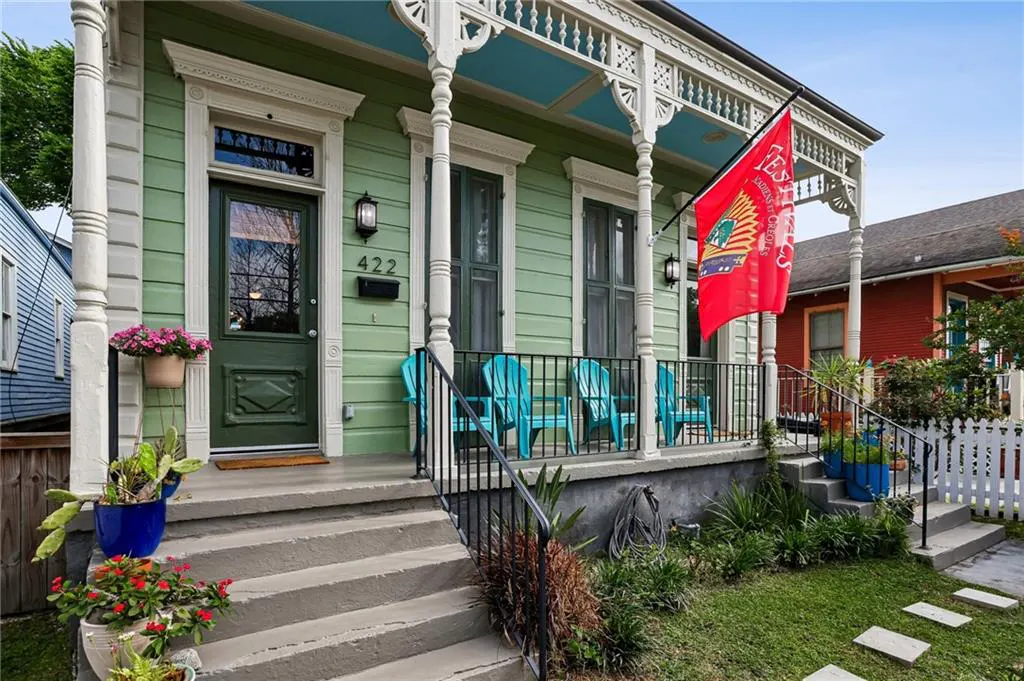 Two-story green house with a porch, blue chairs, and a red flag. The porch has white columns and a blue ceiling.