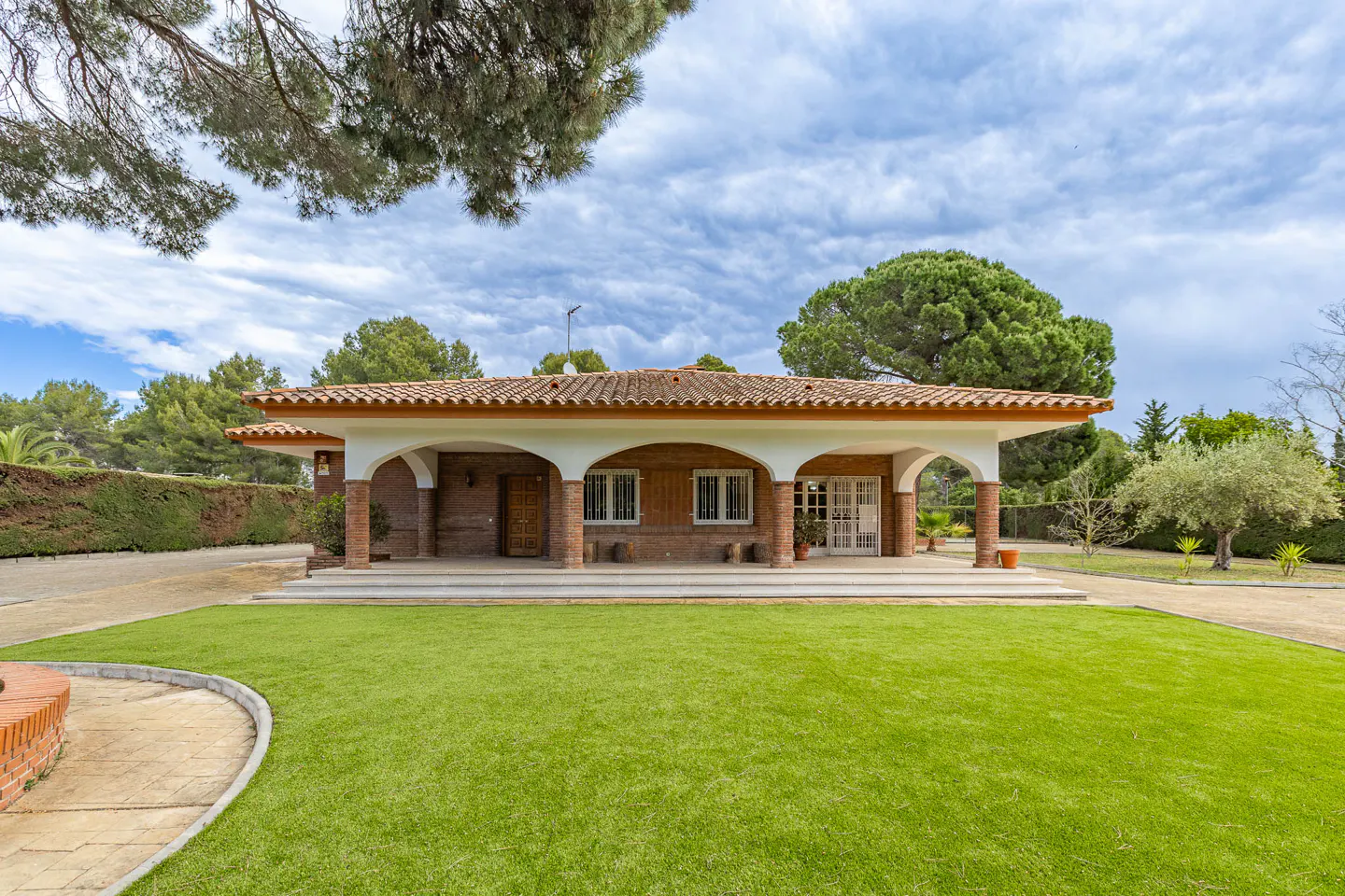 Single-story house with a red tile roof, brick columns, and a large green lawn under a cloudy sky.