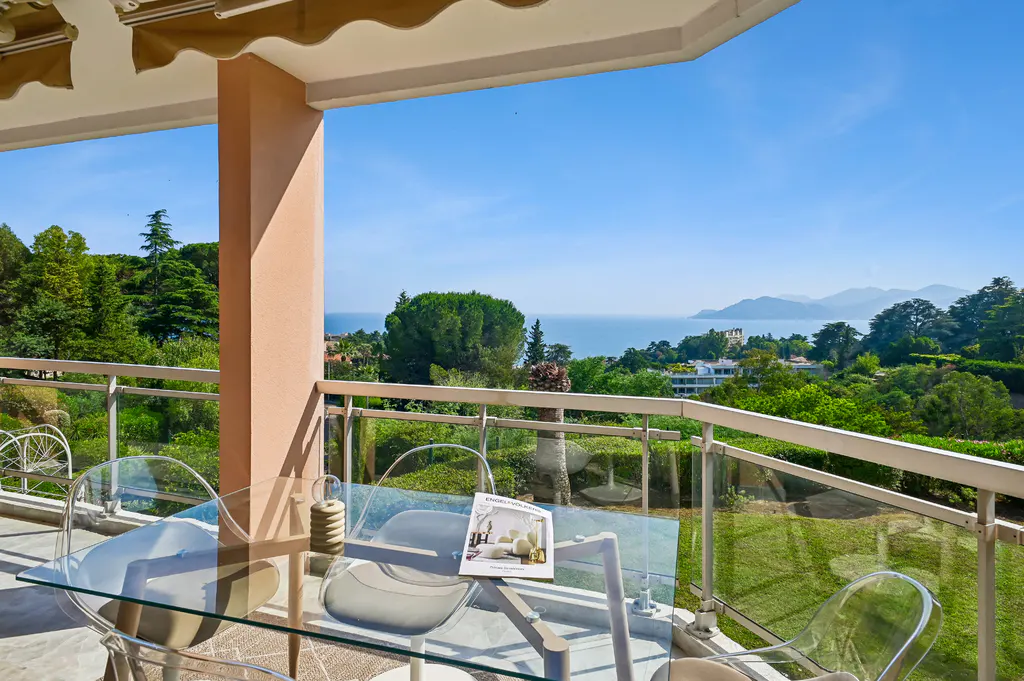 Glass dining table on a balcony overlooking lush green trees, a coastal landscape, and a clear blue sky.