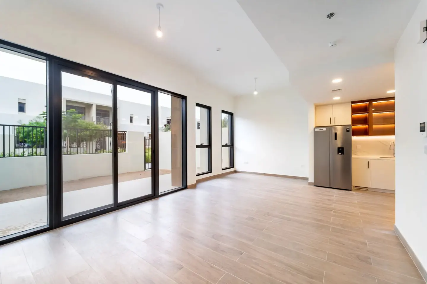 Bright, empty living space with tile floors, white walls, and black-framed sliding glass doors to a patio. Kitchenette with stainless steel fridge.