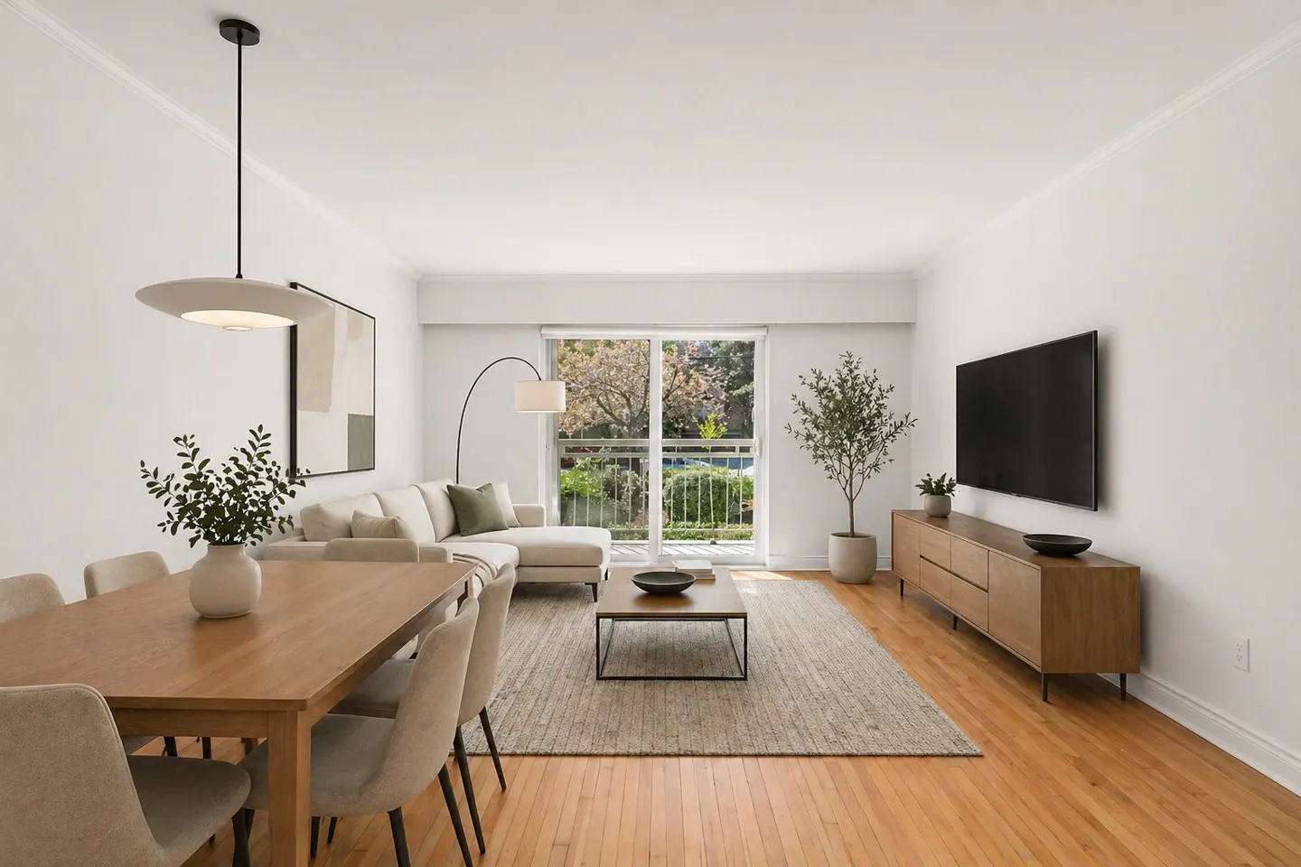 Bright living room with wood floors, white walls, and a dining table. A beige sectional sofa faces a TV, with a balcony visible through sliding glass doors.